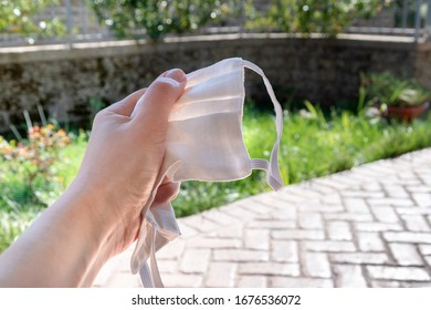 Female Hand Squeezes, Crumples, Holds A Protective Mask In The Garden On A Background Of Green Grass And A Fence. The Concept Of Quarantine Fatigue And Home Isolation