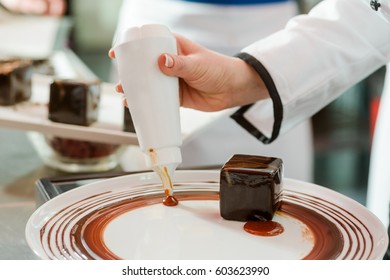 Female Hand Squeezing A Bottle With Berry Sauce, Decorating A Dessert Plate With A Cake On It, Close-up. French Pastry Class.