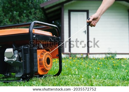 Female hand starts a portable electric generator standing on the grass in front of a summer house in summer evening