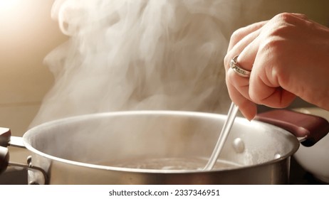 Female hand stirring boiling soup in metal pan with spoon. Vapour slowly rising from hot water - Powered by Shutterstock