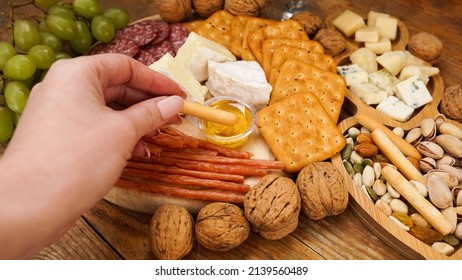 Female Hand Takes A Breadstick And Dips It In Honey. Cheese And Meat Platter With Assorted Cheeses, Crackers, Nuts On Wooden Background