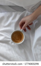 female hand takes cup of coffee in the morning, slow and lazy breakfast in bed on white bedding in the hotel room. copy space