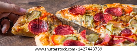 female hand takes a slice of Pepperoni Pizza with Mozzarella cheese, salami, bacon, Tomato sauce, pepper, Spices and pickled cucumbers. Italian pizza on wooden table background