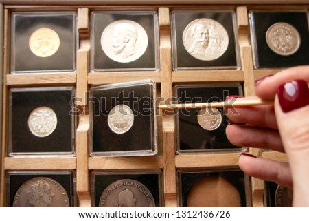Female hand taking out a metal coin in a transparent plastic protection square capsule from a wooden display case with numismatic collection with a wooden stick. Coin holder case for numismatist.