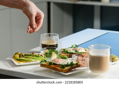 Female Hand Throw Sesame Seeds On A Sliced Avocado. Fried Egg With Toast, Parsley, Sliced Avocado, Butter On A White Plate. Morning Breakfast. Top View.