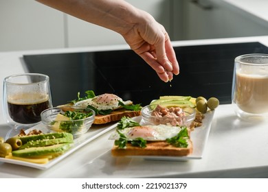 Female Hand Throw Sesame Seeds On A Sliced Avocado. Fried Egg With Toast, Parsley, Sliced Avocado, Butter On A White Plate. Morning Breakfast. Top View.