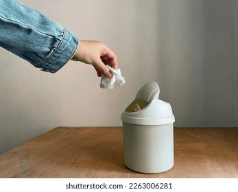 Female hand throwing away a crumpled tissue in small dustbin above wooden table