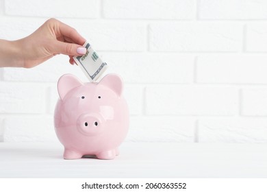 Female hand throwing cash in pink piggybank on white brick wall background