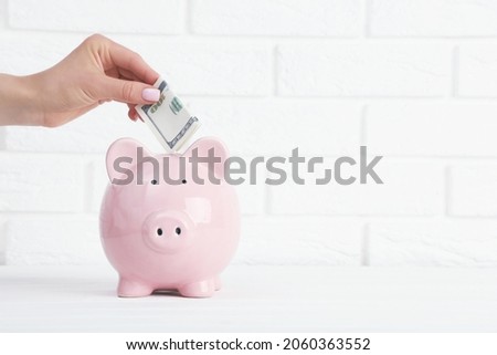 Female hand throwing cash in pink piggybank on white brick wall background
