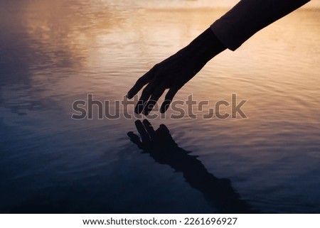 Female hand touching the calm ocean lake water surface reflecting a beautiful colorful summer sunset
