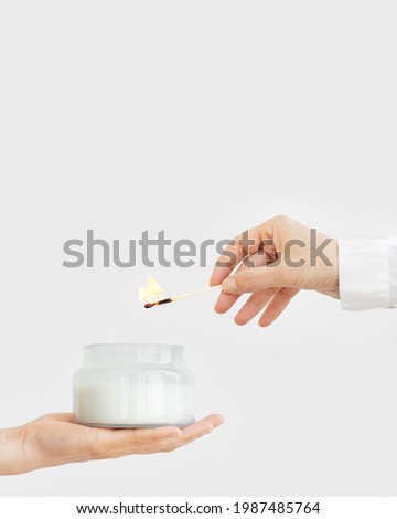 Female hand uses matchstick to light fragrant candle in glass jar with natural ingredients on light background. Natural candles for pleasant moments and relaxation at home.