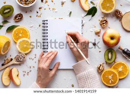 Female hand write in notebook on healthy food background, women diet nutrition recipe menu, fresh summer fruit granola seeds on white table organic super food, health care detox, top view, copy space