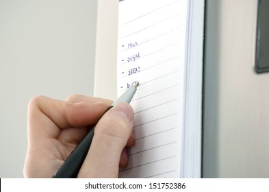 Female Hand Writing Grocery List On A Sticky Reminder Note On A Refrigerator.