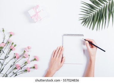 Female hand writing in a notebook at the desk, top view. On the white table lay flowers and a box with a gift and ribbon. - Powered by Shutterstock