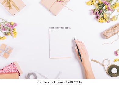 Female Hand Writing In A Paper Notebook On A White Table With Decorations, Top View