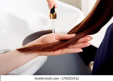 Female hands applying oil on long womans brown hair. Hair care cosmetics, bath beauty spa products. Female hair serum for treatment hair tips against beauty salon interior, bathroom