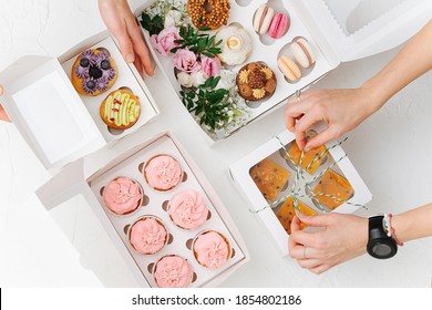 Female hands beautifully and carefully arranging colorful sweets in white windowed boxes. Zefir, cookies, macarons and cakes decorated with branches and flowers. Top view.