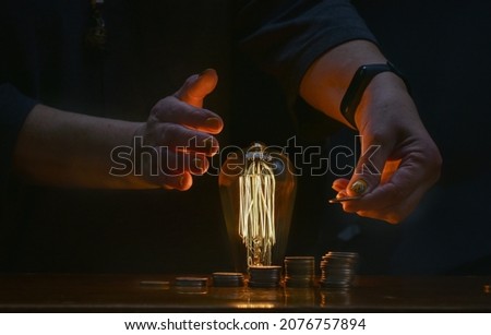 Female hands with a coin, columns of coins in the form of a growing diagram and a burning incandescent lamp on a dark background. The concept of the growth of world energy prices. Selective focus