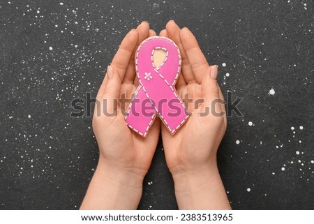 Female hands with cookie in shape of pink ribbon on black grunge background. Breast cancer awareness concept