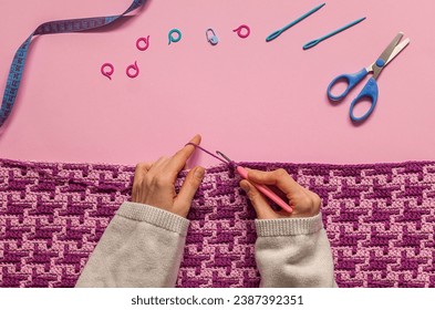 Female hands crocheting a purple mosaic blanket. Crochet fabric and crochet tools on a pink background. Top view. Copy space.