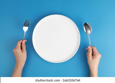 Female hands with cutlery and empty plate on blue background. Meal preparation concept. Top view