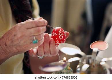 Female Hands Of An Elderly Lady Painting A Red Easter Egg With A Traditional White Design Pattern