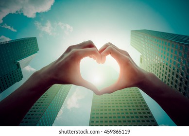 Female Hands In The Form Of Heart Against The Sunshine On Sky With Modern Business Building. Hands In Shape Of Love Heart - Love Business Concept.