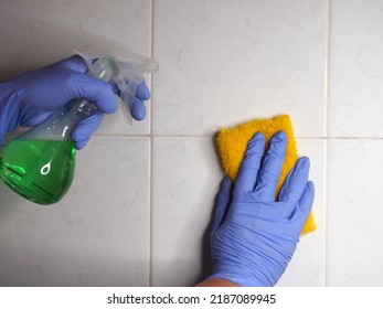 Female Hands In Gloves Hold A Yellow Sponge And A Spray Bottle With Detergent Against The Background Of A White Tiled Wall In The Bathroom.