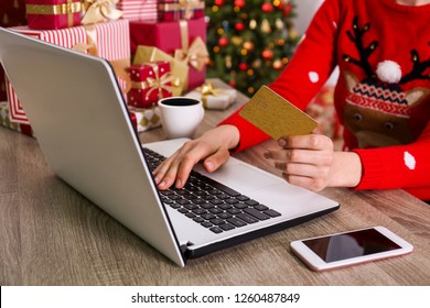 Female hands with golden credit card, laptop, christmas presents, decorated tree on background. Woman picking presents online, eshopping on her laptop. Wooden table, copy space, close up.