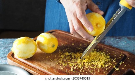 Female hands grating lemon with stainless steel zester or grater over wooden cutting board on rustic blue table with fresh lemons. Healthy eating, Food ingredient preparing theme. (selective focus)