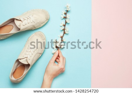 Female hands hold beige fashionable espadrilles with spring flowering branch on pastel blue pink background. Creative concept of spring shoes, fashion blog or magazine concept Flat lay top view