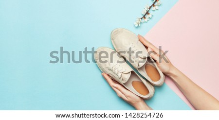 Female hands hold beige fashionable espadrilles with spring flowering branch on pastel blue pink background. Creative concept of spring shoes, fashion blog or magazine concept Flat lay top view
