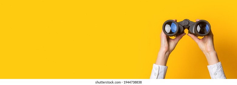 female hands hold black binoculars on a bright yellow background. Banner.