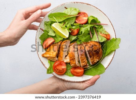 Female hands hold a bowl with salad of chicken breast, fillet with lettuce and cherry tomatoes on a plate on a light background. The concept of healthy, nutritious nutrition.