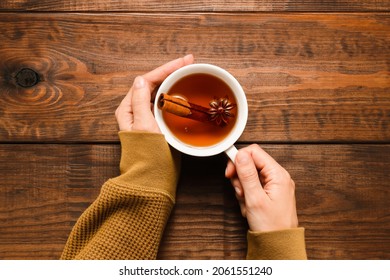 Female hands hold cup of tea with spices, anise and cinnamon. Autumn and winter drink top view on wooden table background