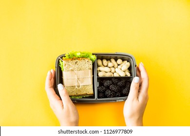 Female Hands Hold Lunchbox With Food - Sandwich, Nuts And Berries On A Yellow Background. Top View, Flat Lay, 