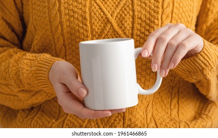 Female hands hold mock up white empty mug, cup for your design and logo close-up. Woman in yellow knitted sweater autumn winter. Blank template for promotional text message or promotional content