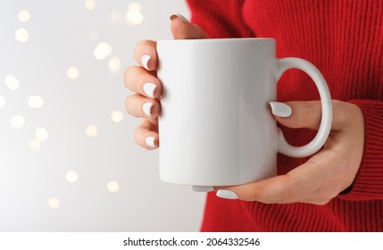 Female Hands Hold A Mockup White Tea Mug In A Red Winter Sweater, Gold Bokeh. For Your Logo And Design. Christmas New Year Concept. Template, Empty