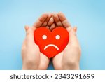 female hands hold a paper heart with a sad smiley face isolated over blue background. broken heart love rejection. hidden narcissist psychology concept