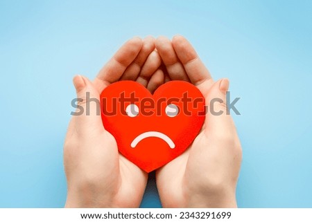 female hands hold a paper heart with a sad smiley face isolated over blue background. broken heart love rejection. hidden narcissist psychology concept Stockfoto © 