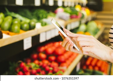 Female hands hold the phone and check the shopping list in the store against the background of vegetables and fruits