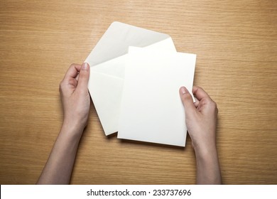 A Female Hands Hold(grip) A White Envelope And Postcard On The Wooden Desk, Top View At The Studio.