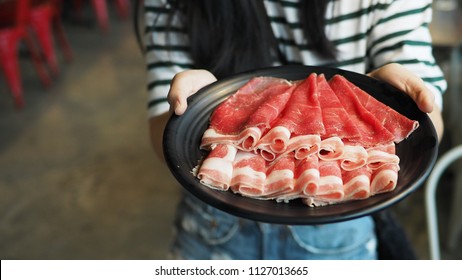 Female hands holding black plate of sliced beef for yakiniku or shabu shabu hot pot. Woman in casual striped t-shirt serving one portion of various meats. (close up, selective focus, space for text)