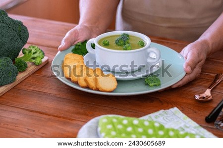 Female hands holding a bowl with handmade broccoli soup and croutons, fresh raw green broccoli on table.H ealthy nutrition concept, eat vegetables, vegetarian, vegan cuisine.