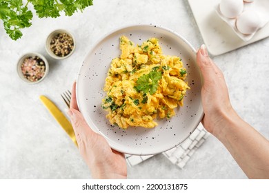 Female hands holding breakfast scrambled eggs with green herbs, parsley in white plate on grey neutral table, golden fork and knife, top view