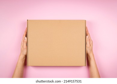 Female Hands Holding Brown Cardboard Box On Pastel Pink Background. Top View To Mockup Parcel Box. Packaging, Shopping, Delivery Concept