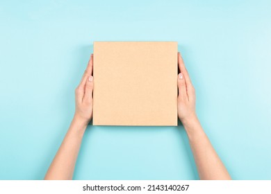 Female hands holding brown ecological package box made of natural corrugated cardboard. Mockup parcel box on light blue background. Top view. Packaging, shopping, delivery concept