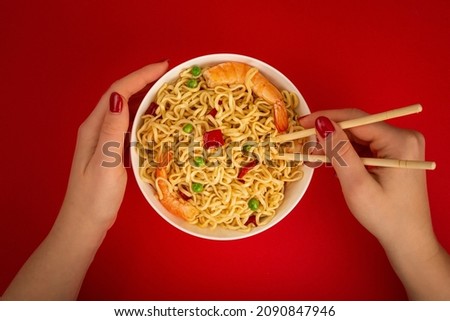 Female hands holding chopsticks and white bowl of asian Chinese noodles top view flat lay with shrimps and vegetables on red minimal paper background, minimal foodphoto concept