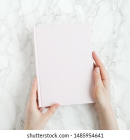 Female hands holding closed book with blank cover on light background. Copy space. Top view