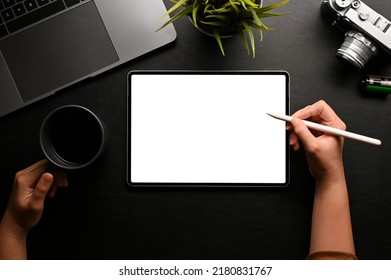A female hands holding a coffee cup and stylus pen, using digital tablet on her modern black office desk. Tablet white screen mockup. top view